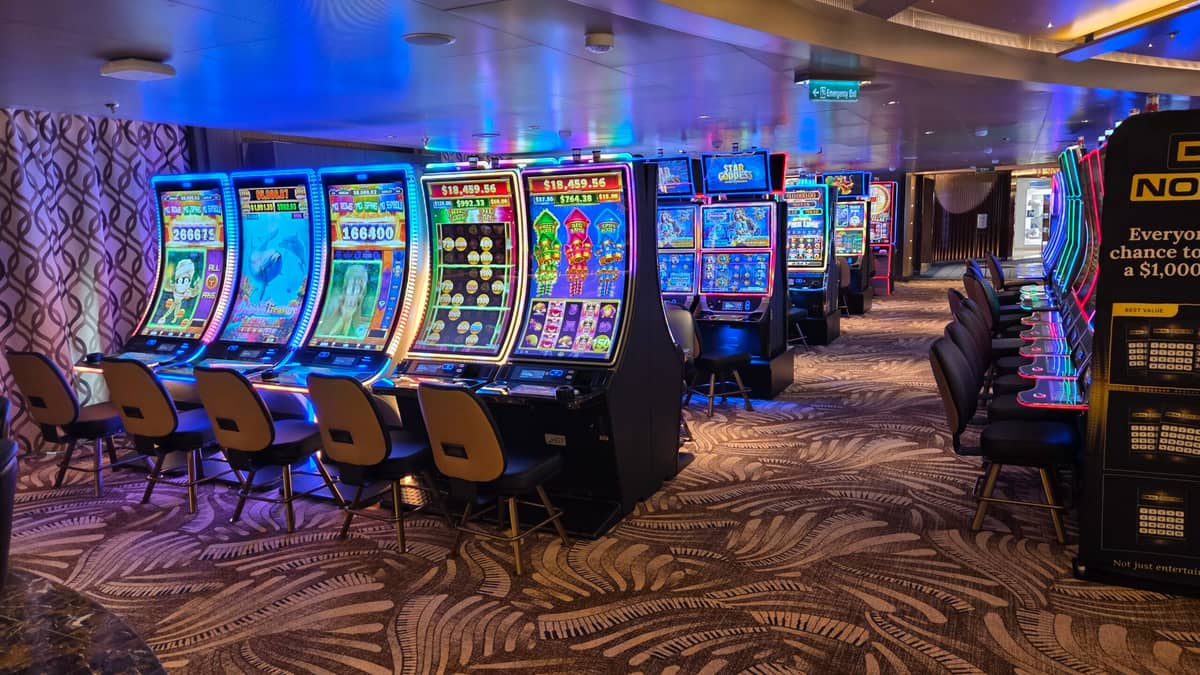 A row of brightly lit slot machines lines one side of a casino floor, with patterned carpet and empty chairs in front of each machine. The area appears well-maintained and inviting.