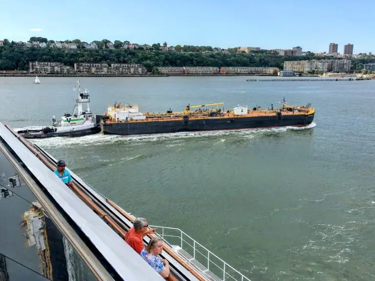 A tugboat pulls a barge along a river, passing near a large cruise lines vessel. Two people lean on the railing of the vessel while buildings and trees line the opposite riverbank under a clear sky.