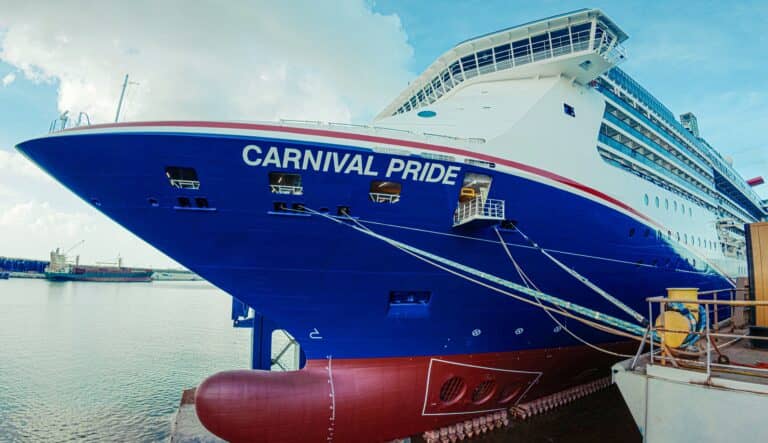 A large cruise ship named Carnival Pride is docked at a port, with ropes securing it to the pier. The ship's blue and white hull is visible under a partly cloudy sky.
