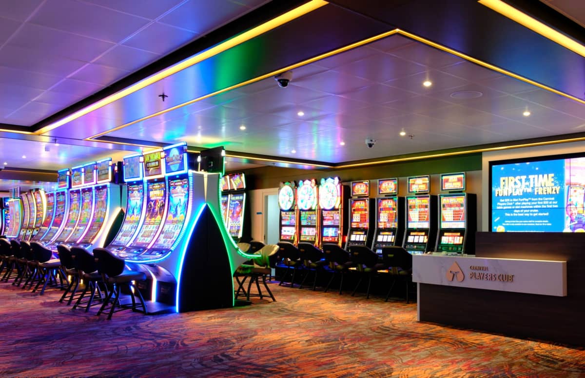 A brightly lit casino interior features rows of slot machines, colorful neon lights, and a carpeted floor. An unoccupied reception desk with a sign reading “Players Club” sits in the foreground.