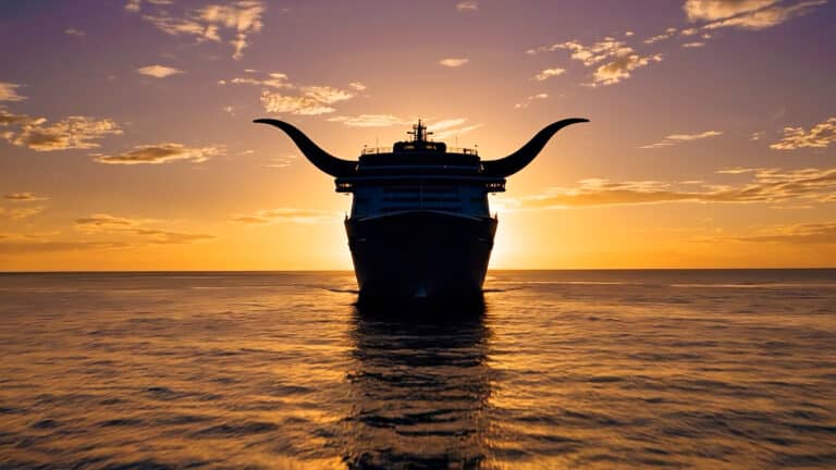 A large Carnival Cruise Line ship with horn-shaped whale tail structures on its upper deck is silhouetted against an orange and purple sunset sky, reflecting on calm ocean water.