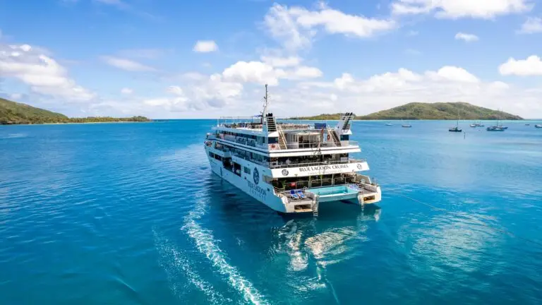 Large white and blue Blue Lagoon Cruises ship anchored in clear blue water, with small islands and boats visible in the distance under a partly cloudy sky.