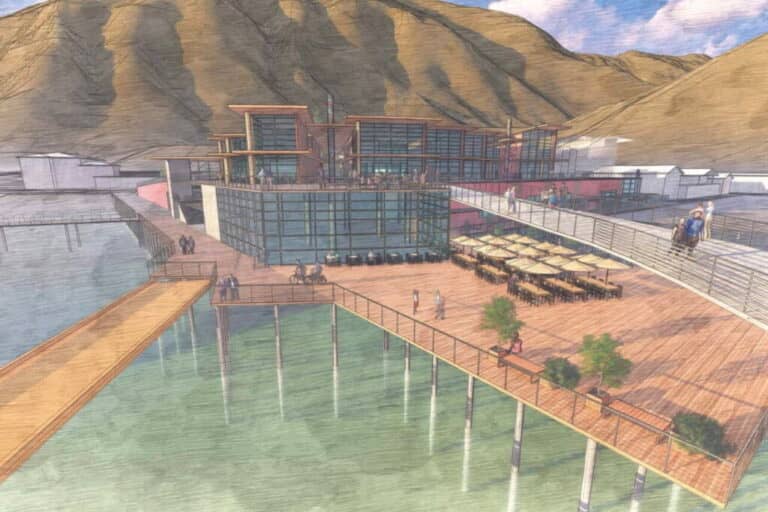 A rendering of a modern waterfront building with large glass windows, outdoor seating with umbrellas, and a boardwalk on stilts extending over calm water, set against a backdrop of hills.