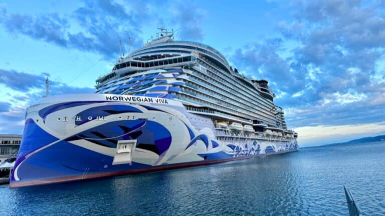 A large cruise ship named Norwegian Viva is docked at a port under a partly cloudy sky. The vessel features blue and white wave patterns on its hull and multiple decks with balconies.