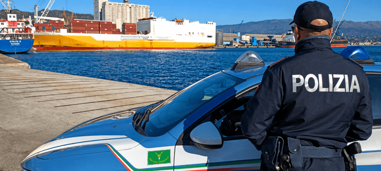 A police officer wearing a "Polizia" jacket stands next to a police car by a port with shipping containers, a yellow and white cargo ship, and industrial buildings in the background.
