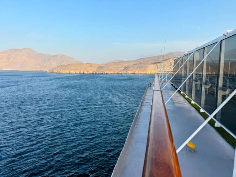 The image shows the deck of a cruise ship with clear glass railings, sailing near a rocky coastline under a blue sky. The calm sea and shoreline stretch across the background, reminiscent of cruises navigating near the Strait of Hormuz.