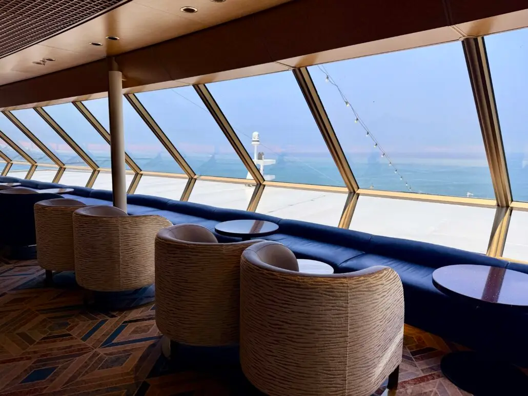 Rows of cushioned chairs and round tables are arranged by large slanted windows, overlooking the ocean on a cruise ship—an inviting setting featured in many Volendam Review highlights. The area is empty, with lights strung outside above the deck.