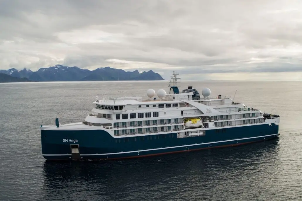 A blue and white cruise ship named SH Vega sails on calm water with mountainous terrain visible in the distance under a cloudy sky.