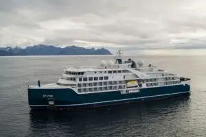 A blue and white cruise ship named SH Vega sails on calm water with mountainous terrain visible in the distance under a cloudy sky.