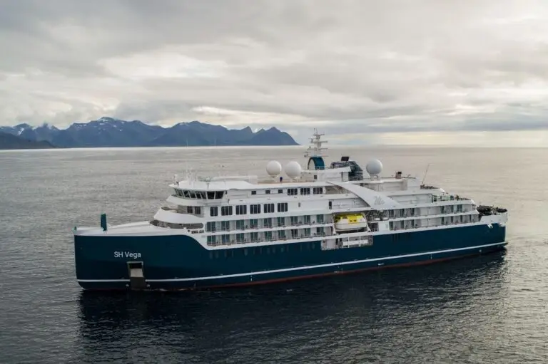 A blue and white cruise ship named SH Vega sails on calm water with mountainous terrain visible in the distance under a cloudy sky.