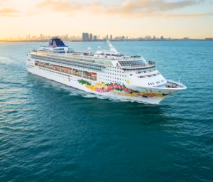 The NCL Norwegian Sky, adorned with colorful hull artwork, sails on calm blue water near a city skyline at sunset. The sky is partly cloudy with soft sunlight, marking the ship's final season in this stunning setting.