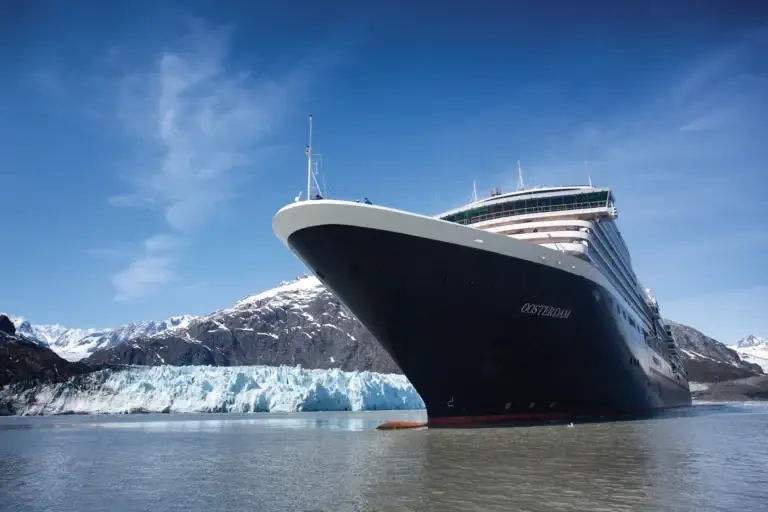 A large Holland America Line cruise ship, featuring recent ship upgrades, is anchored on calm water near a glacier with snow-capped mountains in the background under a clear blue sky.