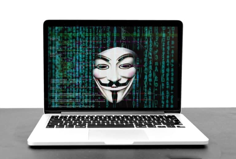 A laptop displays a screen with green code and a large Guy Fawkes mask, symbolizing hacking or anonymous activity—evoking events like the Carnival Data Breach. The laptop sits on a black surface with a white background.