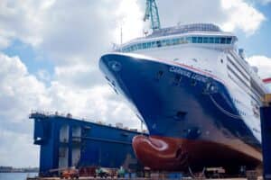 A large Carnival Cruise Ship named "Carnival Legend" is docked at a shipyard, receiving cruise ship updates and a new look, with scaffolding and cranes visible nearby under a partly cloudy sky.