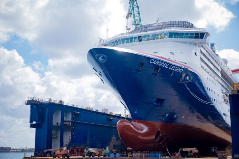 A large Carnival Cruise Ship named "Carnival Legend" is docked at a shipyard, receiving cruise ship updates and a new look, with scaffolding and cranes visible nearby under a partly cloudy sky.