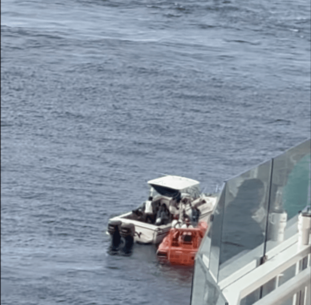 A small white boat and an orange vessel are next to each other on the water near the side of Carnival Mardi Gras, where several people are visible. The calm ocean fills the background during this latest sailing that rescued nine people.
