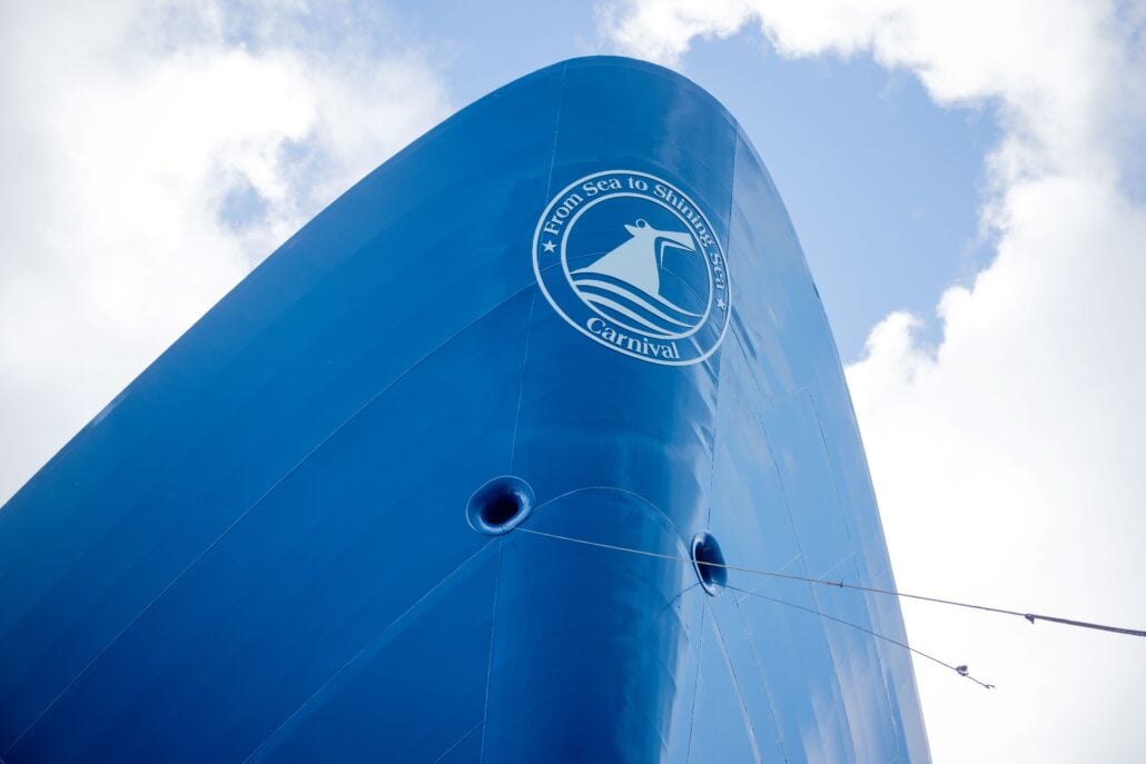Close-up view of the bow of the blue Carnival Legend cruise ship, featuring the line's logo and slogan "From Sea to Shining Sea," set against a partly cloudy sky.