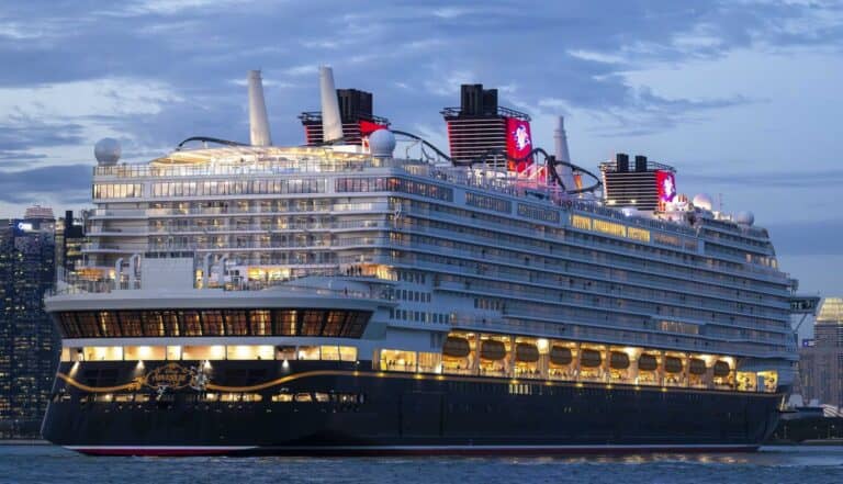 A large cruise ship with multiple decks and illuminated windows is docked in a city harbor at dusk, with buildings visible in the background and a partly cloudy sky overhead.