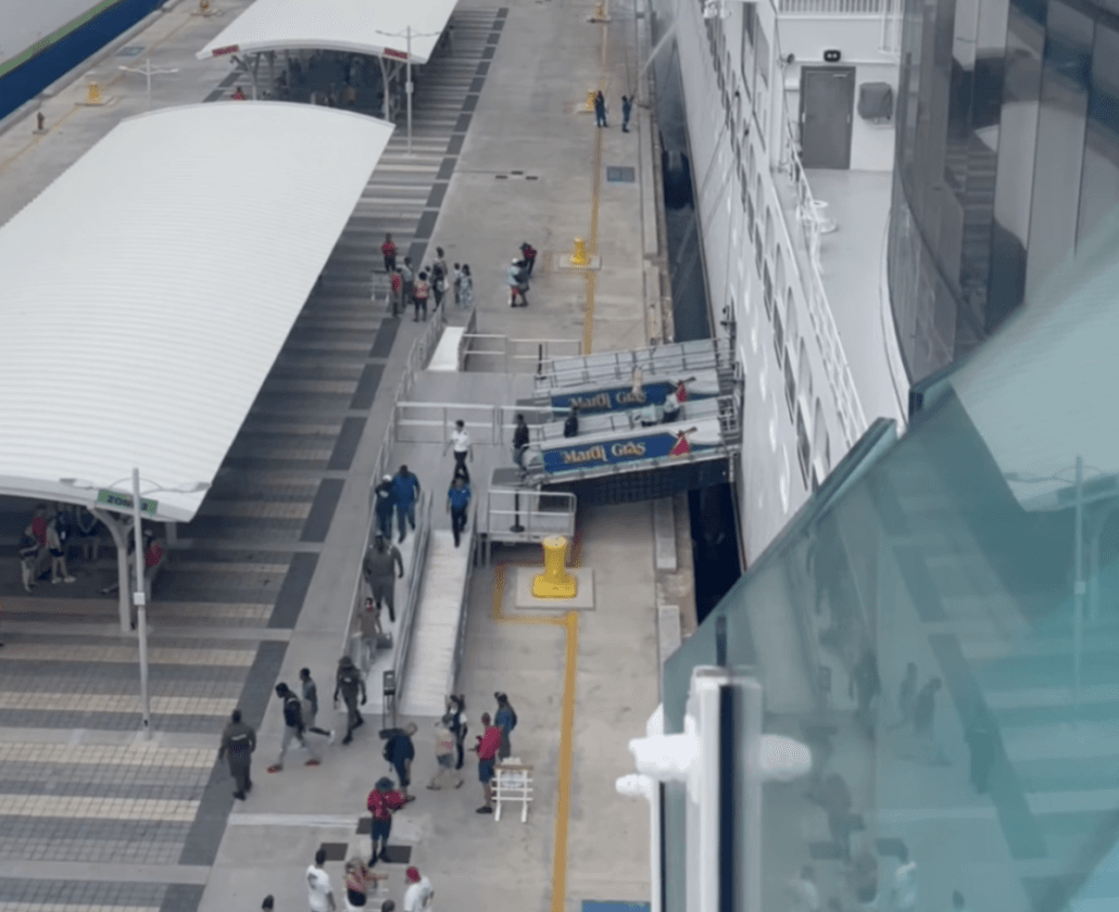 A view from above shows people walking on a cruise terminal dock beside the large white Carnival Mardi Gras ship. Two covered walkways and boarding ramps labeled “Mardi Gras” connect the dock to this latest sailing.