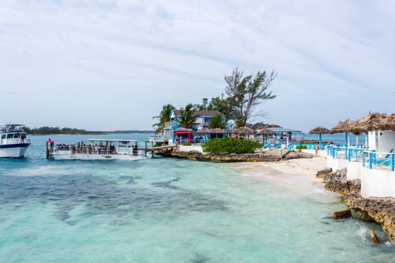A small dock with Carnival passengers boarding boats sits near a sandy beach with clear turquoise water, thatched umbrellas, and white buildings with blue trim under a partly cloudy sky—a perfect scene for a Bahamas excursion.
