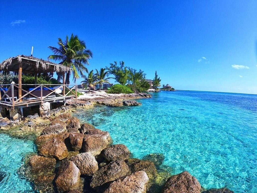 A wooden hut with a thatched roof sits by a rocky shoreline, surrounded by palm trees and clear turquoise water under a bright blue sky—the perfect spot for a relaxing Bahamas excursion.