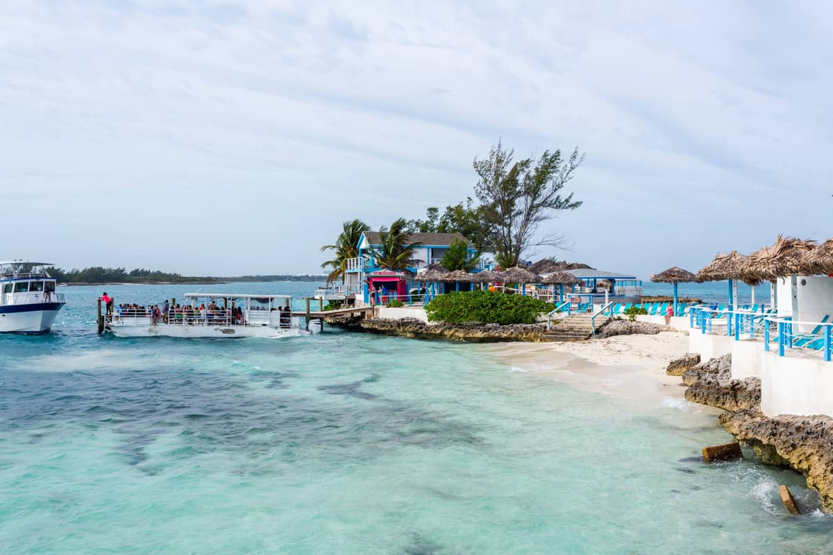 A small dock with Carnival passengers boarding boats sits near a sandy beach with clear turquoise water, thatched umbrellas, and white buildings with blue trim under a partly cloudy sky—a perfect scene for a Bahamas excursion.