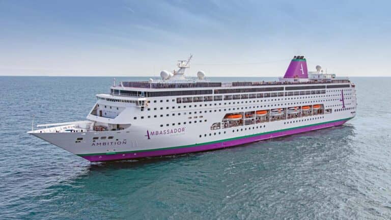 A large white cruise ship named Ambition, operated by Ambassador, sails on calm blue water near France under a clear sky. The ship has multiple decks, orange lifeboats, and a purple funnel with a white "A" logo.