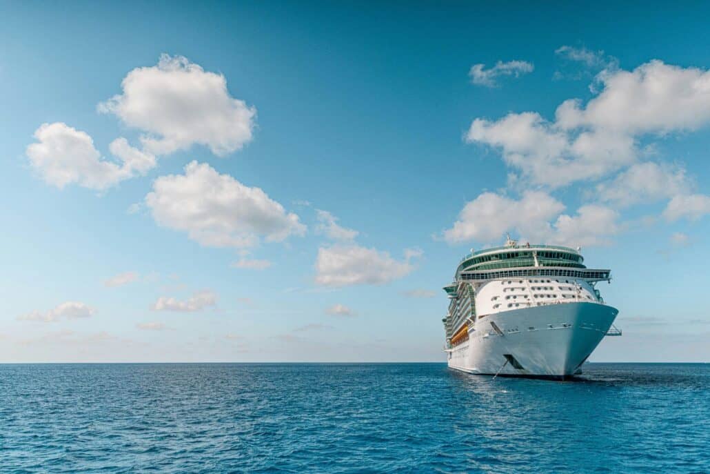 A large white cruise ship is sailing on calm blue ocean water under a clear sky with scattered clouds.