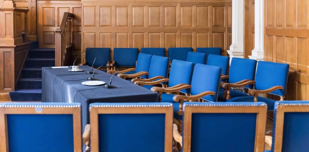 A row of empty blue-cushioned chairs with wooden frames is arranged in a wood-paneled room, likely a jury box in an Italian court. Two tables with microphones and plates are set in front of the chairs.