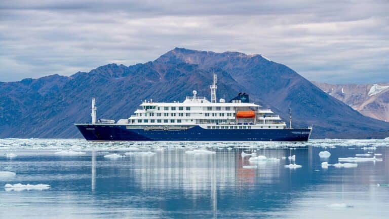 A large cruise ship with a dark blue hull and white upper decks sails through icy Arctic waters, surrounded by floating ice, with rugged mountains in the background under a cloudy sky.