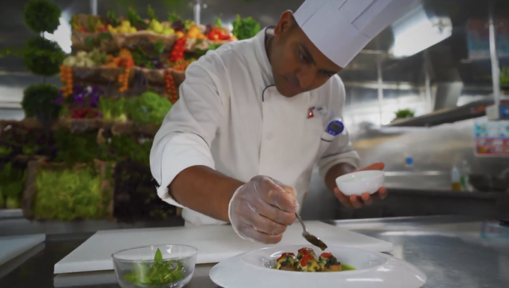 A chef wearing a white uniform and hat carefully garnishes a plated dish in a kitchen. Fresh vegetables are visible in the background, hinting at new menus inspired by Carnival Cruise Line, with a bowl of greens on the counter nearby.