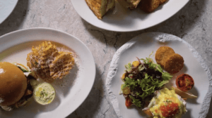 Overhead view of three plates of food on a light stone surface, showcasing dishes inspired by new menus from Carnival Cruise Line: a burger with waffle fries, toast, and a salad with greens, poached eggs, croquettes, and roasted tomato.