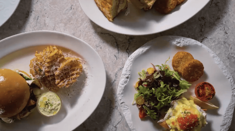 Overhead view of three plates of food on a light stone surface, showcasing dishes inspired by new menus from Carnival Cruise Line: a burger with waffle fries, toast, and a salad with greens, poached eggs, croquettes, and roasted tomato.