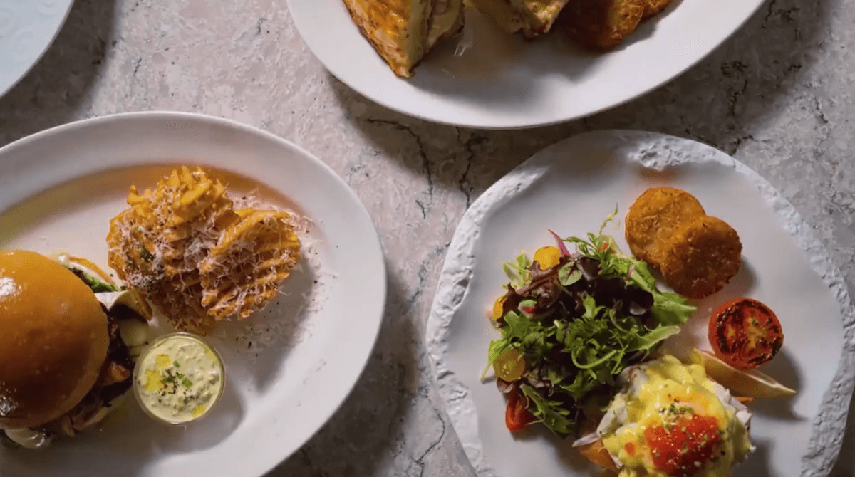 Overhead view of three plates of food on a light stone surface, showcasing dishes inspired by new menus from Carnival Cruise Line: a burger with waffle fries, toast, and a salad with greens, poached eggs, croquettes, and roasted tomato.