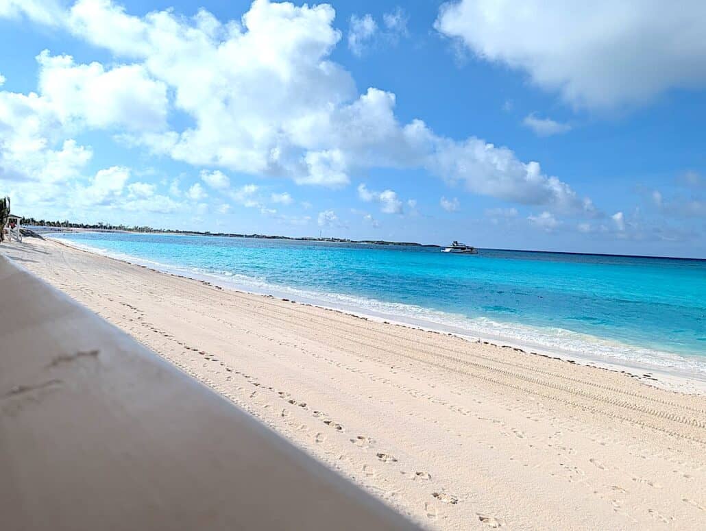 A sandy beach at Half Moon Cay with clear turquoise water under a blue sky dotted with clouds. Faint footprints and tire tracks line the sand as you relax away, watching a small boat floating near the horizon.