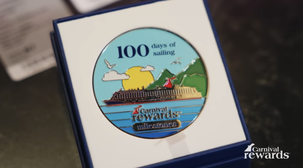A round Carnival Rewards milestones medallion in a display box, featuring an illustration of a cruise ship, mountains, ocean, sun, seagulls, and text reading “100 days of sailing.” Includes a brief Carnival Rewards explanation.