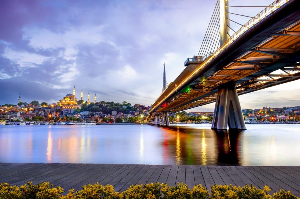 A modern suspension bridge spans over a calm river at sunset near Istanbul Port, with a cityscape and a large mosque featuring multiple minarets in the background. Clouds drift across the sky as soft evening light sets the scene for an unforgettable Yacht Day.