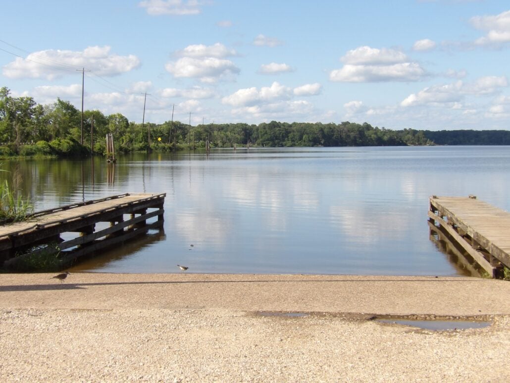 A calm lake with a concrete boat ramp in the foreground, flanked by two wooden docks, invites those who love boating, surrounded by trees and power lines under a blue sky with scattered clouds.