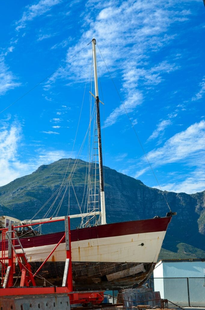 A sailboat with a red and white hull is positioned on a dry dock for maintenance, an ideal scene for any Long Distance Boat Transport planning guide. Metal supports hold the boat, set against green mountains and a blue sky with scattered clouds.