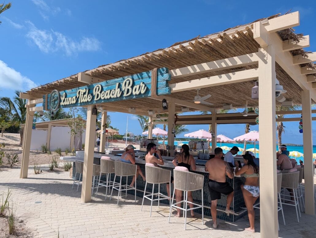 People relax away at Suna Tide Beach Bar under a wooden pergola with a thatched roof on Half Moon Cay. Pink umbrellas and the ocean create a vibrant backdrop for this sunny Carnival getaway.