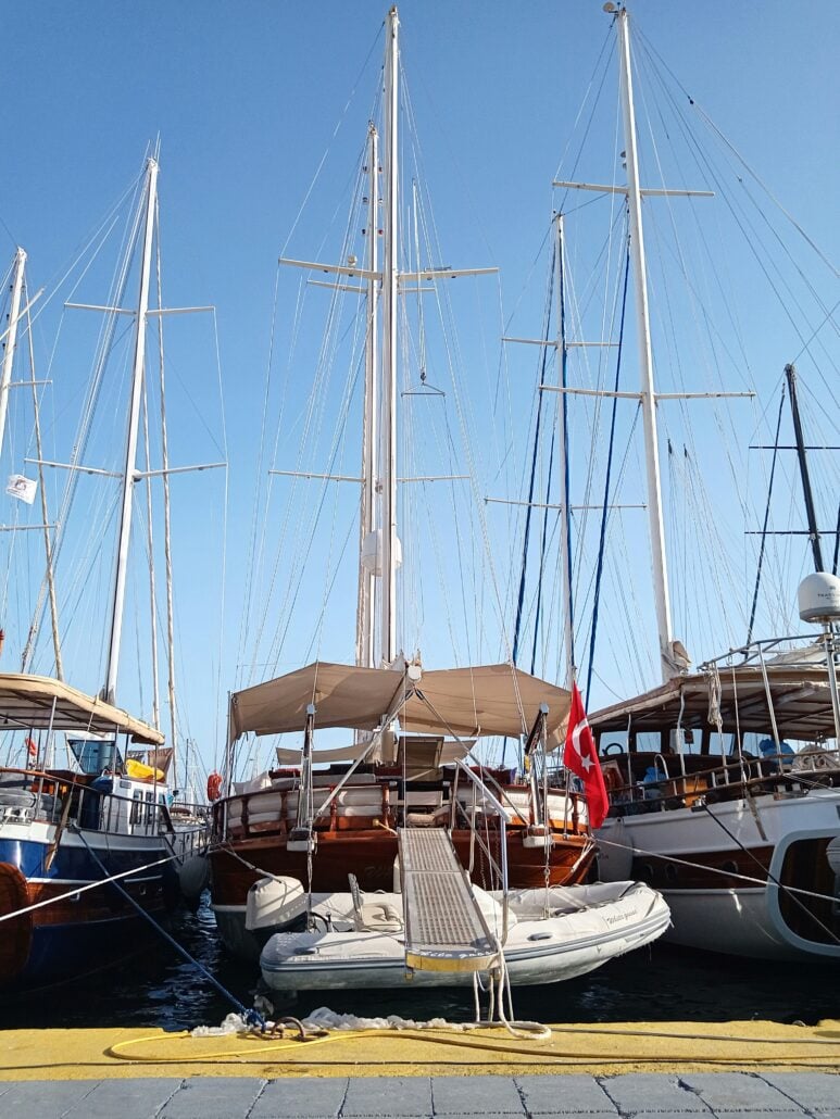 Several sailboats and yachts are docked side by side at a marina under a clear blue sky. Perfect for seasonal travel, a ramp leads from the dock to one boat displaying a Turkish flag, while an inflatable dinghy is moored in front.