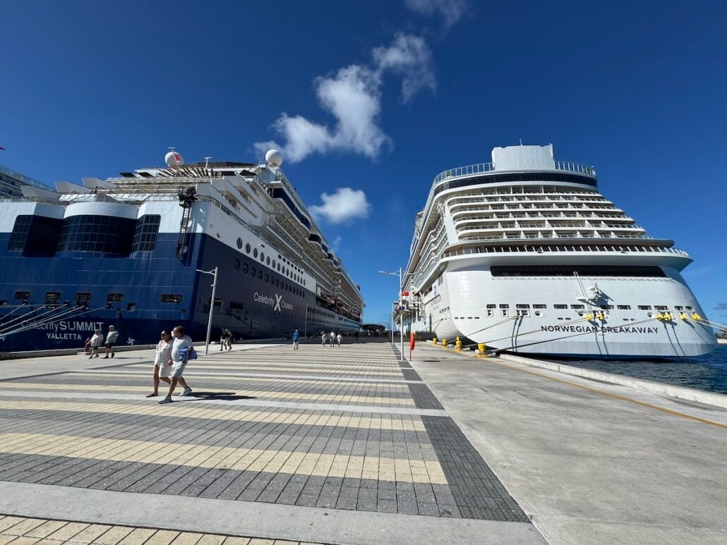 Two large cruise ships from major cruise lines are docked side by side at a port under a clear blue sky. Several people are walking along the wide, tiled promenade, unaware of recent fights that led to some passengers being banned-for-life.