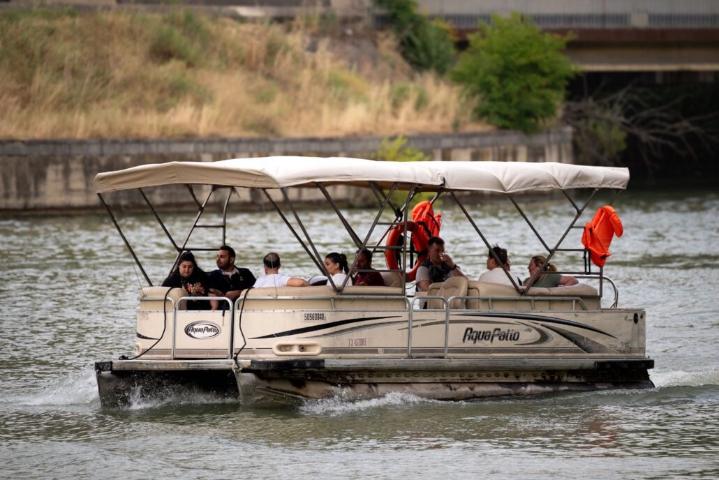 A beige pontoon boat with a canopy carries ten people on a river. Some passengers are seated while others stand. Orange life jackets hang on the canopy frame. The background shows a grassy bank and a concrete structure.