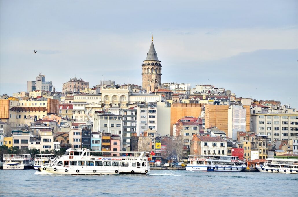 A view of Istanbul’s shoreline with several boats on the water near the Istanbul port and the historic Galata Tower rising among densely packed buildings under a partly cloudy sky.
