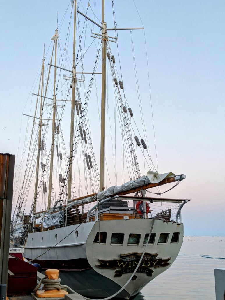 A large white sailing ship with multiple masts and rigging is docked at a pier, perfect for seasonal boat travel. The ship’s stern displays the name “Windy” along with “Chicago.” The sky is clear with pastel colors at sunset or sunrise.