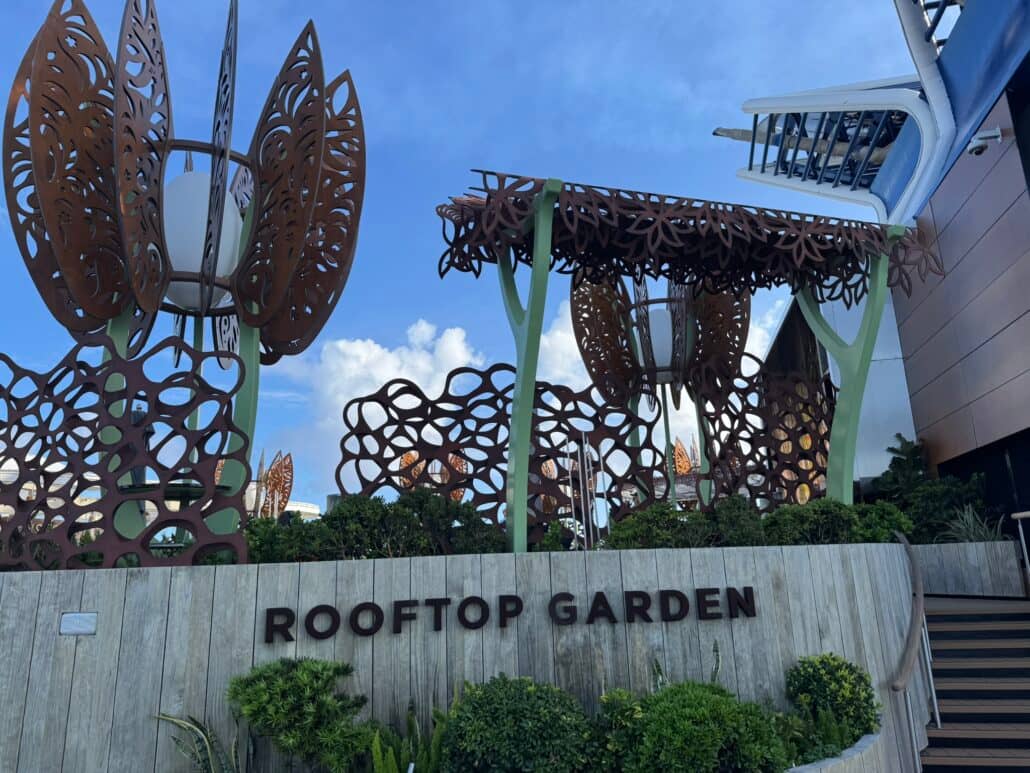 A rooftop garden with sculptural metal structures, modern wood fencing, and lush green plants under a partly cloudy blue sky—an inviting retreat reminiscent of the serene outdoor spaces found on Celebrity Cruises. The words “Rooftop Garden” are displayed on the fence, evoking a sense of relaxation without the need to consider details like daily gratuity.