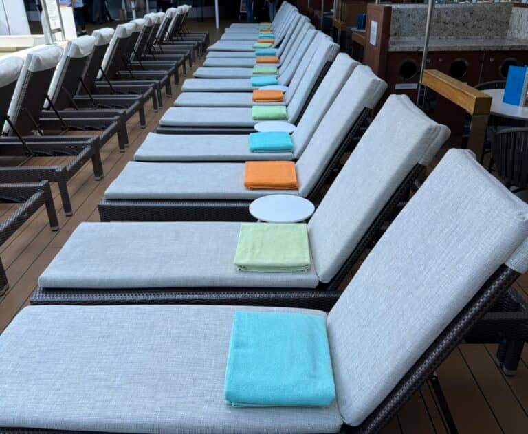 A row of empty deck chairs with light cushions, each with a colored towel—blue, green, or orange—folded at the foot. A white plate rests on one chair. The scene appears to be at a pool or deck area, evoking the tranquil atmosphere of a cruiser in a modern work of fiction.