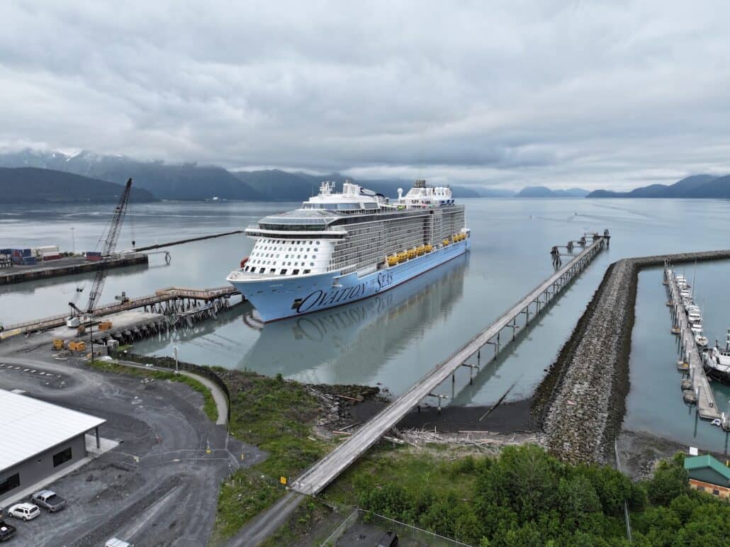 A large cruise ship named “Ovation of the Seas” is docked at a pier in a quiet harbor, surrounded by calm water, mountains, and cloudy skies. A crane and buildings are visible near the shore.