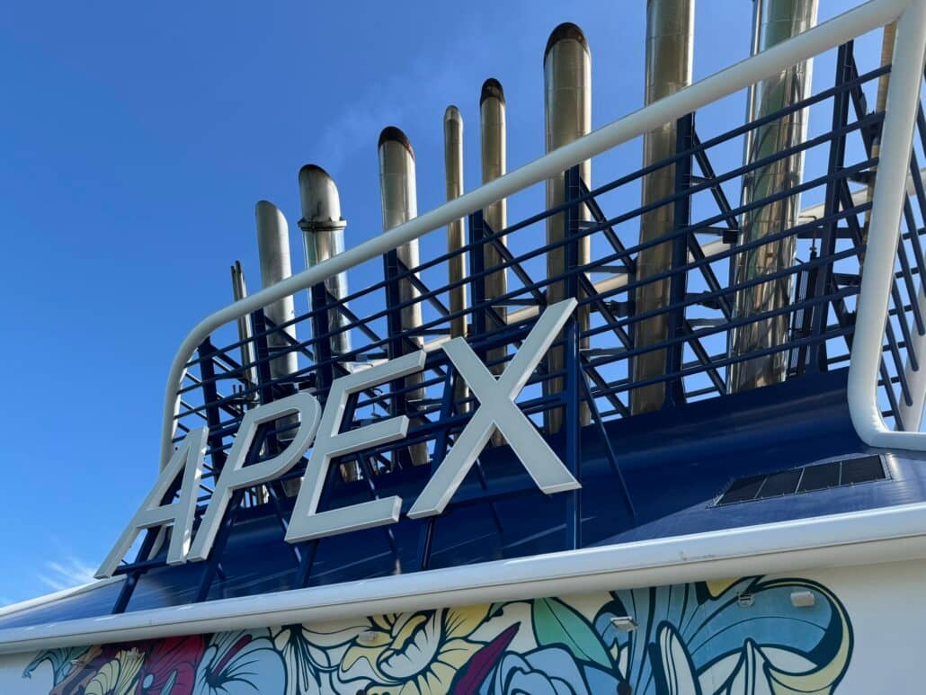 Large white letters spelling "APEX" are mounted on a blue metal structure with multiple exhaust pipes above. A section of colorful mural art is visible at the bottom, and the sky is clear and blue, capturing a vibrant moment aboard Celebrity Cruises where details like the daily gratuity add to your overall experience.