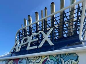 Large white letters spelling "APEX" are mounted on a blue metal structure with multiple exhaust pipes above. A section of colorful mural art is visible at the bottom, and the sky is clear and blue, capturing a vibrant moment aboard Celebrity Cruises where details like the daily gratuity add to your overall experience.